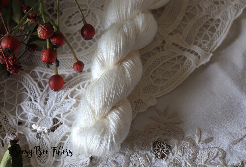 Puede incluir: Un ovillo de hilo de seda blanco, presentado sobre un encaje blanco con el texto "Beesly Bee Fibers" en la esquina inferior izquierda.