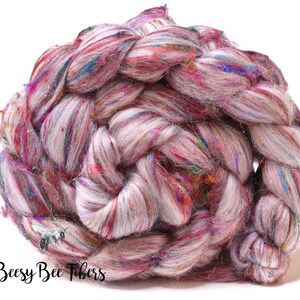 May include: A braided skein of hand-spun yarn in shades of pink, purple, and white, with colorful flecks of blue, green, and gold. The yarn is thick and textured, perfect for knitting or crocheting projects. The text "Beesy Bee Fibers" is visible.
