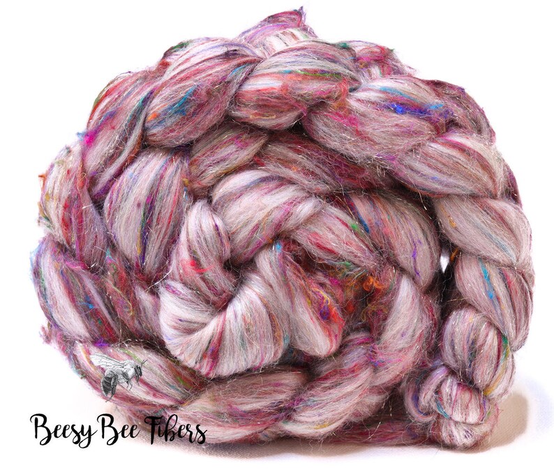 May include: A braided skein of hand-spun yarn in shades of pink, purple, and white, with colorful flecks of blue, green, and gold. The yarn is thick and textured, perfect for knitting or crocheting projects. The text "Beesy Bee Fibers" is visible.