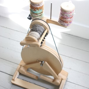May include: A wooden spinning wheel with a large wheel and two bobbins. The bobbins are filled with colorful yarn. The spinning wheel is on a wooden base.
