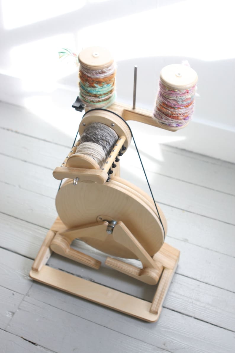 May include: A wooden spinning wheel with a large wheel and two bobbins. The bobbins are filled with colorful yarn. The spinning wheel is on a wooden base.
