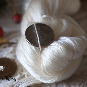 Puede incluir: Una madeja de hilo de seda blanco con una moneda de 25 c&eacute;ntimos encima para mostrar el tama&ntilde;o del hilo. El hilo est&aacute; sobre un encaje blanco.