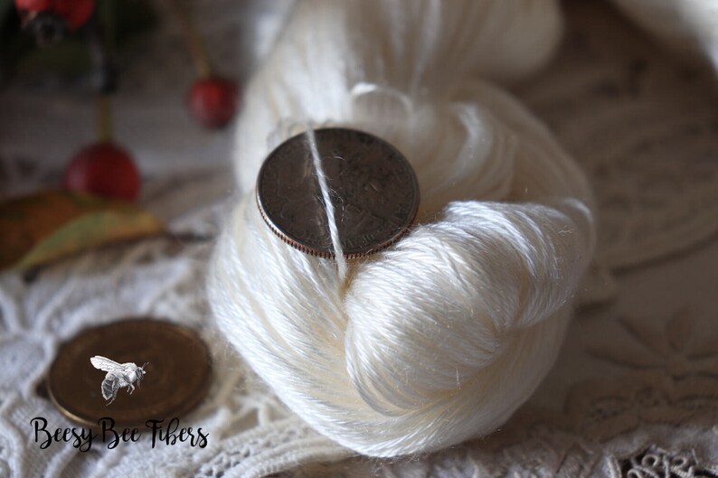 Puede incluir: Una madeja de hilo de seda blanco con una moneda de 25 c&eacute;ntimos encima para mostrar el tama&ntilde;o del hilo. El hilo est&aacute; sobre un encaje blanco.