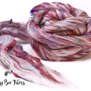 May include: A close-up of a skein of colorful fiber yarn. The yarn is a mix of white, pink, red, and blue, with strands of silver glitter. The yarn is twisted and coiled, with some strands extending outward. The text "Beesy Bee Fibers" is at the bottom.