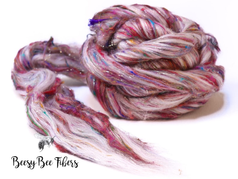 May include: A close-up of a skein of colorful fiber yarn. The yarn is a mix of white, pink, red, and blue, with strands of silver glitter. The yarn is twisted and coiled, with some strands extending outward. The text "Beesy Bee Fibers" is at the bottom.
