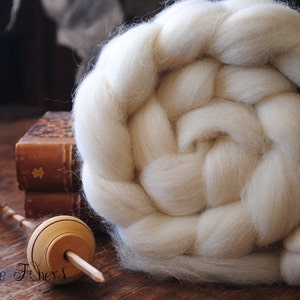 Puede incluir: Un hilado de lana blanco, tambi&eacute;n conocido como top, est&aacute; dispuesto en forma de roseta trenzada. Un torno de madera est&aacute; sobre una superficie de madera.