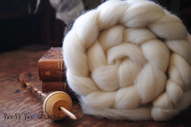 Puede incluir: Un hilado de lana blanco, tambi&eacute;n conocido como top, est&aacute; dispuesto en forma de roseta trenzada. Un torno de madera est&aacute; sobre una superficie de madera.