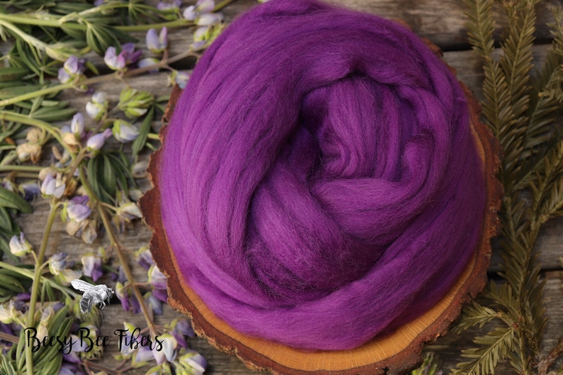 May include: A close-up of a deep purple wool roving, a type of fiber used for spinning yarn or felting. The roving is arranged in a circular shape and is sitting on a wooden surface.