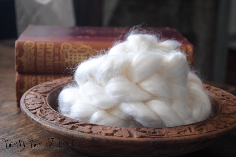 May include: A white, fluffy pile of natural fiber sits in a wooden bowl with intricate carvings. The fiber is arranged in a braided pattern, creating a soft and textured appearance.