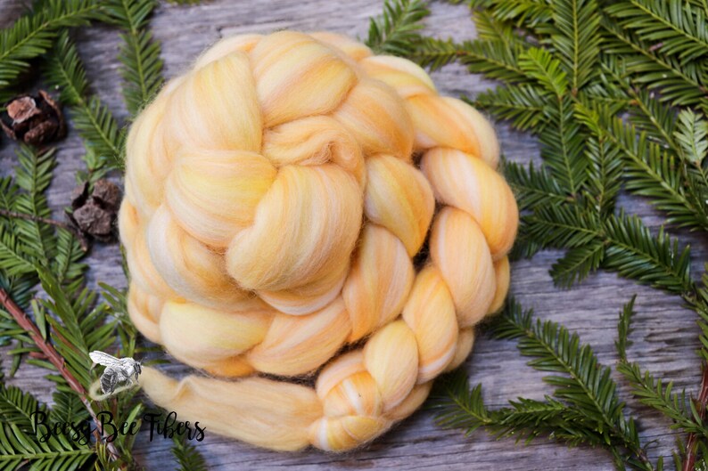 Puede incluir: Un ovillo de lana amarilla y naranja trenzado sobre un fondo blanco. La lana est&aacute; dispuesta en forma circular y est&aacute; rodeada de ramas de pino verdes.