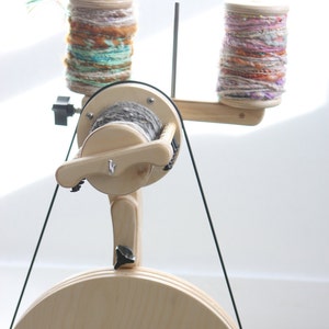 May include: A wooden spinning wheel with a large wooden wheel and two spools of yarn. The yarn is a mix of pink, purple, and white.