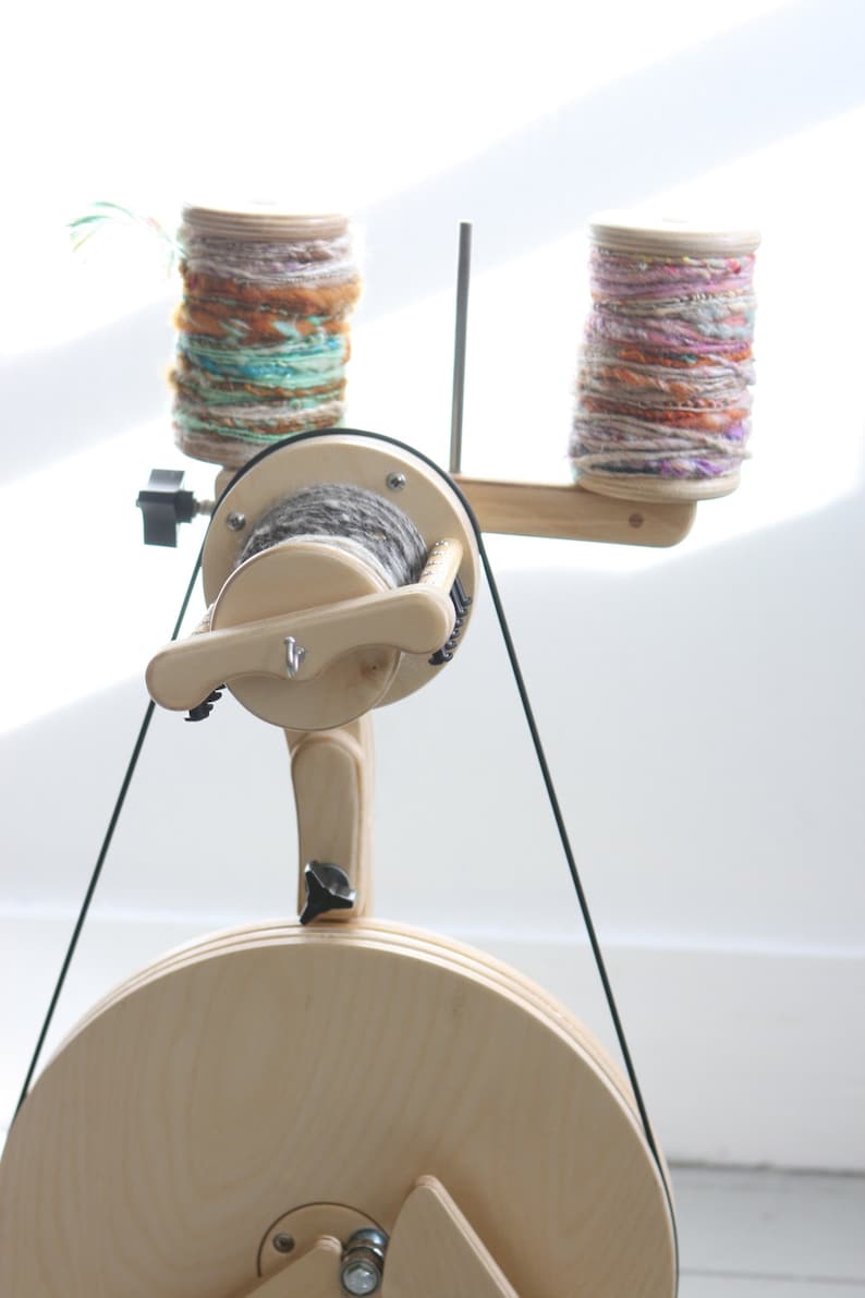 May include: A wooden spinning wheel with a large wooden wheel and two spools of yarn. The yarn is a mix of pink, purple, and white.
