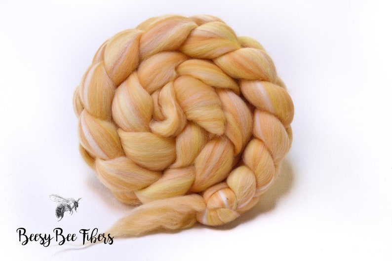 Puede incluir: Una bola trenzada de vell&oacute;n de lana amarilla y naranja con una raya blanca que la atraviesa. El vell&oacute;n est&aacute; hecho por Beesy Bee Fibers.