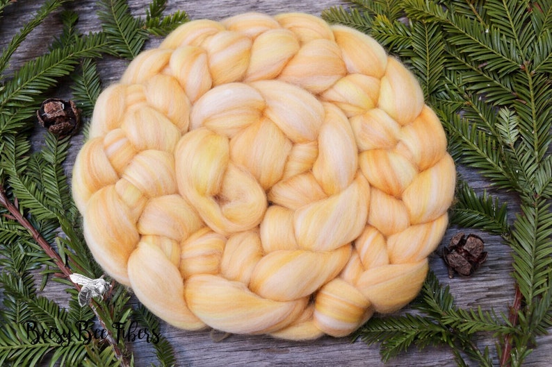 Puede incluir: Un paquete de fibra de alpaca amarilla y blanca trenzada, dispuesto en forma circular. La fibra es suave y esponjosa, y es perfecta para hilar o fieltro.