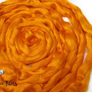 Puede incluir: Primer plano de una fibra de seda naranja brillante, hilada en una forma circular suelta. La fibra es suave y esponjosa, y es perfecta para hilar o fieltro. La imagen tambi&eacute;n incluye el texto "Beesly Bee Fibers".