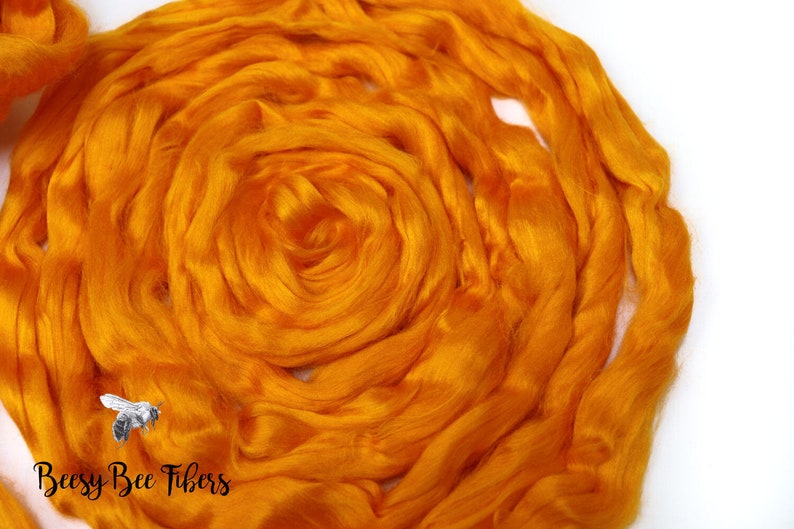 Puede incluir: Primer plano de una fibra de seda naranja brillante, hilada en una forma circular suelta. La fibra es suave y esponjosa, y es perfecta para hilar o fieltro. La imagen tambi&eacute;n incluye el texto "Beesly Bee Fibers".