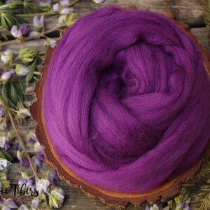 May include: A close-up of a purple wool roving, a type of fiber used for spinning yarn, displayed on a wooden slice. The roving is soft and fluffy, and the color is a rich, deep purple.