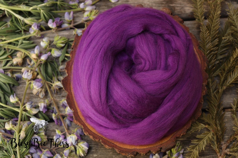 May include: A close-up of a purple wool roving, a type of fiber used for spinning yarn, displayed on a wooden slice. The roving is soft and fluffy, and the color is a rich, deep purple.