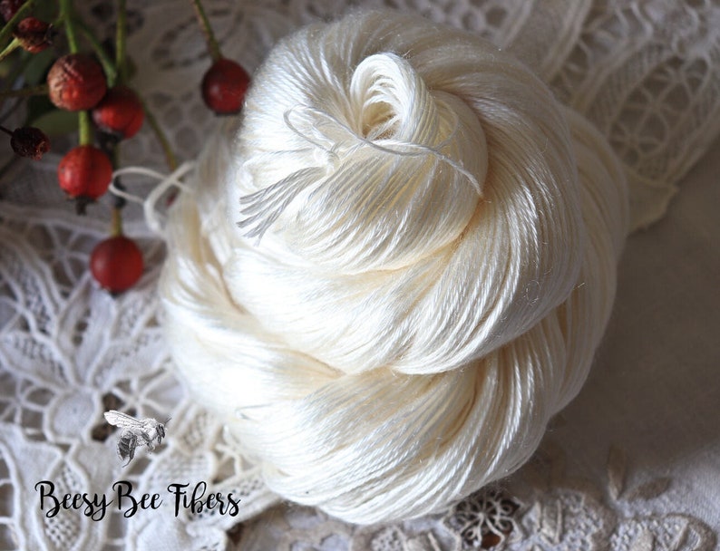 Puede incluir: Una madeja de hilo de seda blanco, enrollada en forma de bola, sobre un fondo de encaje blanco. El hilo es suave y delicado, y la madeja tiene un di&aacute;metro de unos 25 cm.