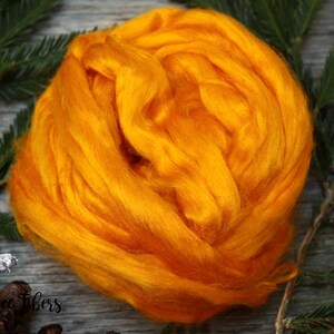 Puede incluir: Primer plano de una fibra esponjosa de color naranja brillante, probablemente seda o lana, dispuesta en una bola suelta. La fibra est&aacute; sobre una superficie de madera con ramas de pino verdes y pi&ntilde;as marrones.