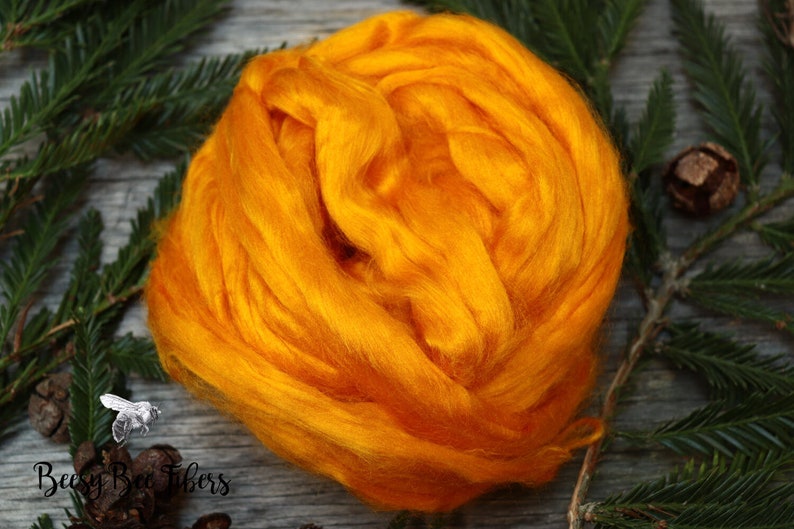 Puede incluir: Primer plano de una fibra esponjosa de color naranja brillante, probablemente seda o lana, dispuesta en una bola suelta. La fibra est&aacute; sobre una superficie de madera con ramas de pino verdes y pi&ntilde;as marrones.