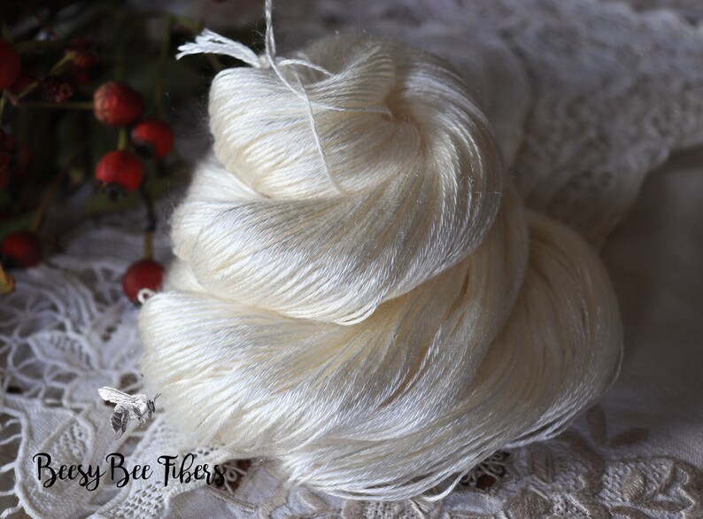 Puede incluir: Un ovillo de hilo de seda blanco, enrollado de forma suelta y listo para tejer o hacer ganchillo. El hilo es suave y delicado, con un brillo sutil.