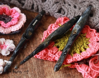 May include: Three black and grey crochet hooks with a marbled effect, lying on a wooden surface with crocheted flowers and a crocheted square in pink, yellow, and green.