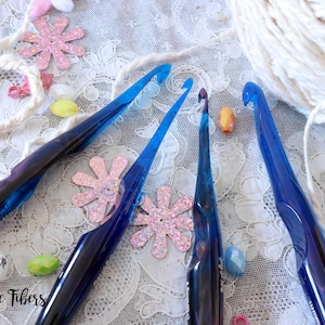 May include: Four blue crochet hooks on a white lace background with colorful embellishments. The hooks are made of plastic and have a shiny finish. The text "Beesly Bee Fibers" is visible in the lower left corner of the image.