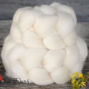 Puede incluir: Una gran trenza de vell&oacute;n de lana blanquecina, ideal para proyectos de arte textil. La imagen muestra un primer plano de la lana suave y texturizada, con un peque&ntilde;o objeto decorativo y flores amarillas en el fondo. El texto "Beesy Bee Fibers" es visible.