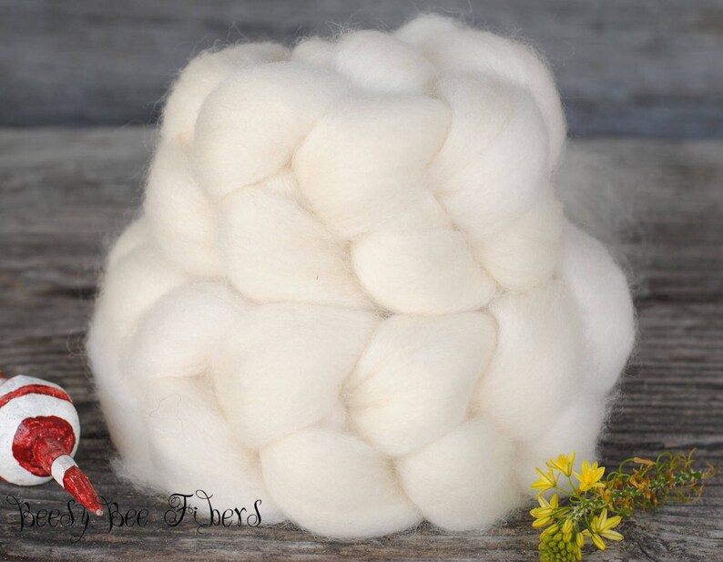 Puede incluir: Una gran trenza de vell&oacute;n de lana blanquecina, ideal para proyectos de arte textil. La imagen muestra un primer plano de la lana suave y texturizada, con un peque&ntilde;o objeto decorativo y flores amarillas en el fondo. El texto "Beesy Bee Fibers" es visible.