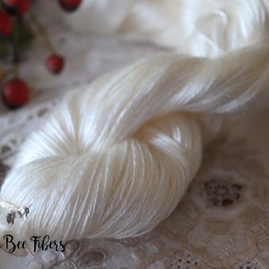 Puede incluir: Una madeja de hilo de seda blanco, con un brillo sutil, se muestra sobre un mantel de encaje blanco. El hilo es suave y delicado, y es perfecto para tejer o hacer ganchillo prendas finas.