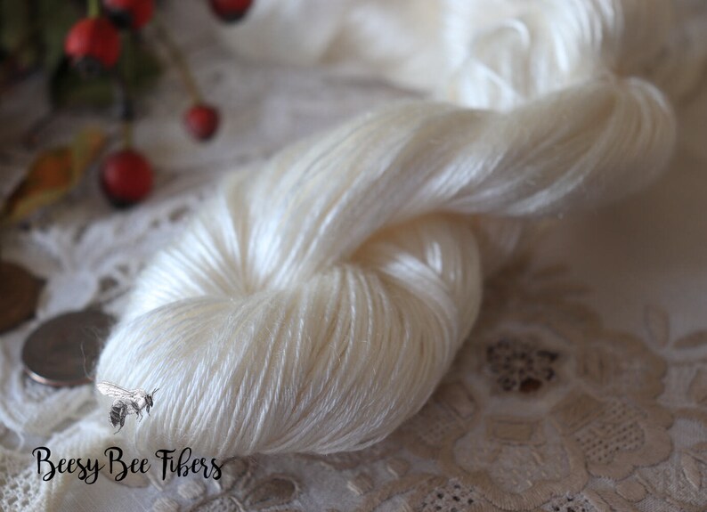 Puede incluir: Una madeja de hilo de seda blanco, con un brillo sutil, se muestra sobre un mantel de encaje blanco. El hilo es suave y delicado, y es perfecto para tejer o hacer ganchillo prendas finas.