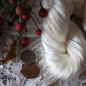 Puede incluir: Una madeja de hilo de seda blanco, mostrada sobre un encaje blanco con bayas rojas y una hoja. El hilo es suave y delicado, y la etiqueta dice "Beesy Bee Fibers".