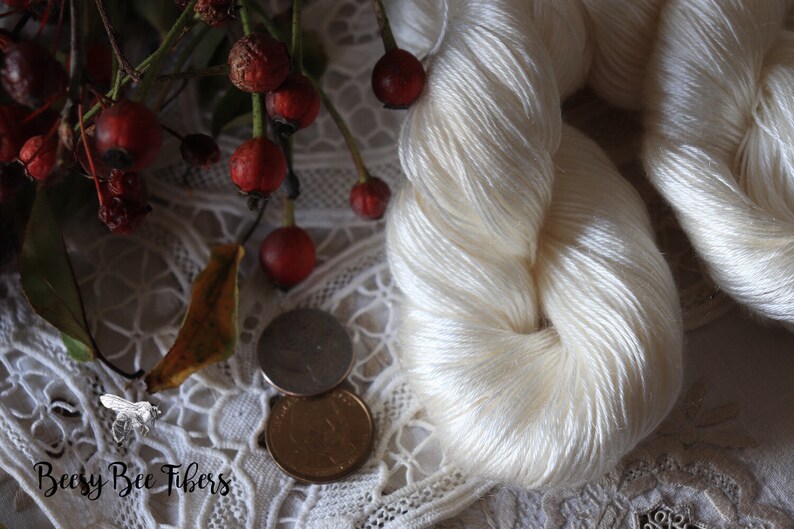 Puede incluir: Una madeja de hilo de seda blanco, mostrada sobre un encaje blanco con bayas rojas y una hoja. El hilo es suave y delicado, y la etiqueta dice "Beesy Bee Fibers".