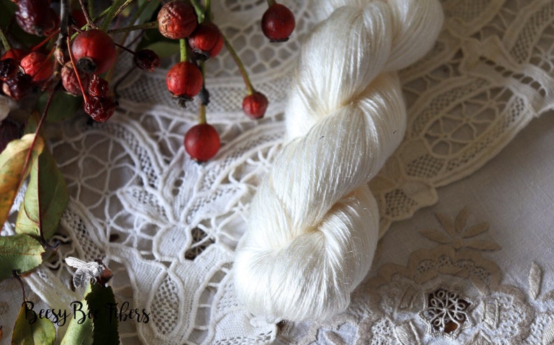 Puede incluir: Una madeja de hilo de seda blanco, con una textura suave y delicada, se muestra sobre un encaje blanco. El hilo est&aacute; retorcido en forma de cuerda, destacando su tacto lujoso.
