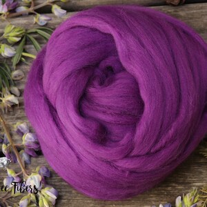 May include: A close-up of a deep purple wool roving, a type of fiber used for spinning yarn or felting. The roving is arranged in a loose, fluffy ball.