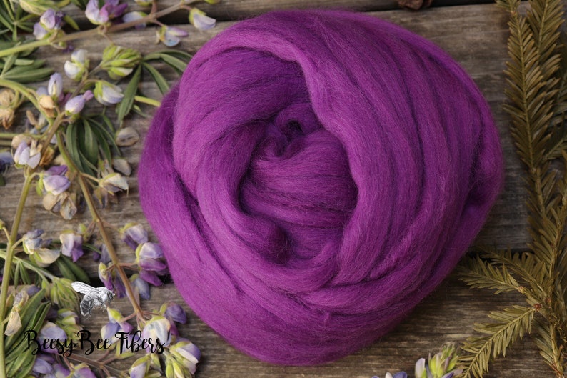 May include: A close-up of a deep purple wool roving, a type of fiber used for spinning yarn or felting. The roving is arranged in a loose, fluffy ball.