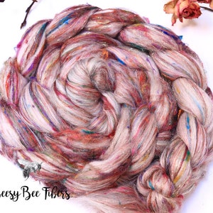 May include: Close-up of a braided skein of hand-spun yarn. The yarn is a mix of light pink, cream, and multicolored fibers with metallic accents. The text "Beesy Bee Fibers" is visible in the lower left corner.