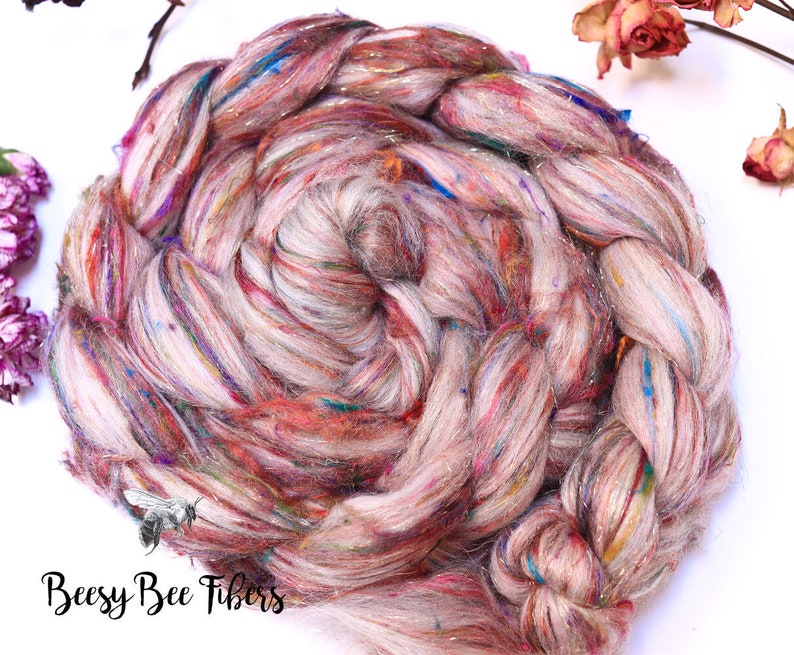 May include: Close-up of a braided skein of hand-spun yarn. The yarn is a mix of light pink, cream, and multicolored fibers with metallic accents. The text "Beesy Bee Fibers" is visible in the lower left corner.