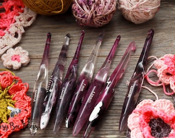 May include: A set of eight purple and clear crochet hooks with different sizes marked on them. The hooks are laid out on a wooden surface with crocheted flowers and yarn balls.