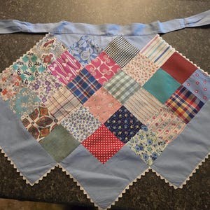 May include: A patchwork apron with a light blue tie and trim. The apron features a diamond pattern of various fabric squares in floral, plaid, and polka dot designs. The trim is white with a scalloped edge.