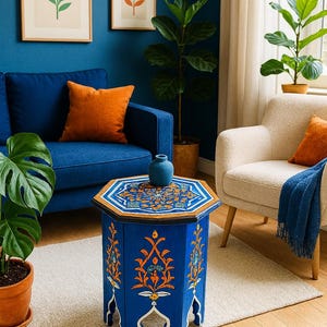 Op de afbeelding: Een blauwe, achthoekige bijzettafel met ingewikkelde oranje en witte bloemmotieven. De tafel heeft een houten blad en poten. Een kleine blauwe vaas staat erop. De kamer heeft een blauwe bank, oranje kussens en ingelijste kunst.