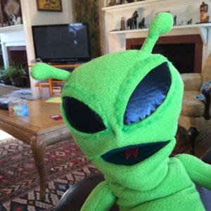 Puede incluir: Marioneta de extraterrestre verde con grandes ojos negros y una boca roja. La marioneta está hecha de tela suave y de felpa y tiene una cabeza larga y puntiaguda.