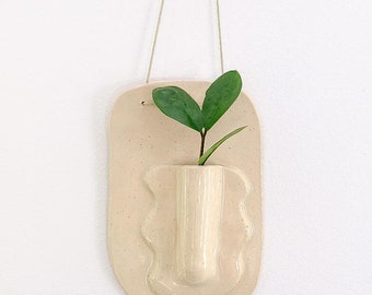 May include: A beige ceramic wall planter with a small vase-like pocket and a green plant. The planter is rectangular with a wavy design around the vase. A beige string is attached for hanging. The background is a white wall.