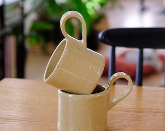 May include: Two beige ceramic mugs stacked on a wooden table. Each mug has a rounded handle and a speckled texture. The mugs are in a neutral colour palette, with a blurred background of greenery and a black chair.