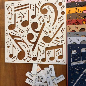 May include: A white stencil with cut-out musical notes and symbols. A selection of coloured stencils are stacked to the right. Small rectangular labels with music genre names such as "Jazz" and "Country" are scattered below.
