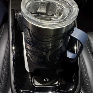 May include: Stainless steel travel mug with a blue handle and a clear lid, resting in a car's center console. The mug has an engraved design and is partially filled. The console is black and has a parking brake symbol.