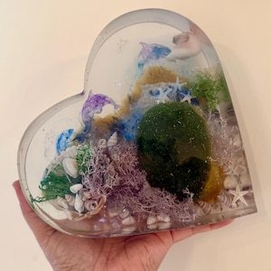 May include: A heart-shaped resin art piece featuring a miniature underwater scene. The clear resin encases a green moss ball, seashells, starfish, and purple and blue accents, creating a unique decorative item.