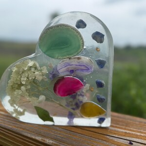 May include: A heart-shaped resin paperweight with embedded gemstones and dried flowers. The clear resin encases green, purple, pink, and yellow stones, along with small blue stones and dried white flowers. The paperweight is displayed on a wooden surface.
