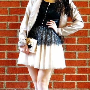 May include: A woman wearing a black leather jacket, a black and white lace skirt, and black heels. She is holding a small, rectangular, cream-colored purse with a gold design. She is standing in front of a brick wall.